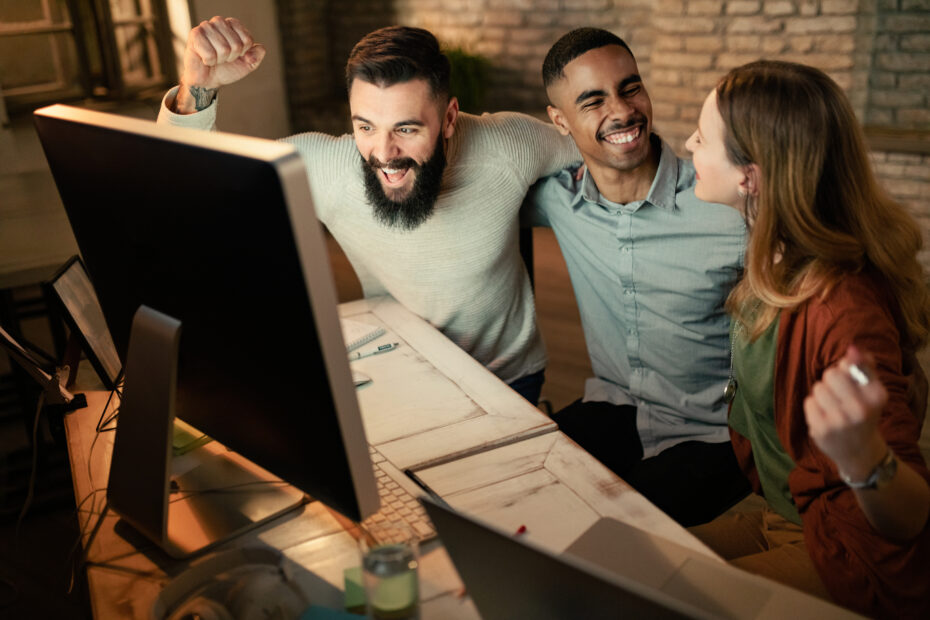 Grupo de colegas felizes comemorando boas notícias em um PC desktop no escritório.